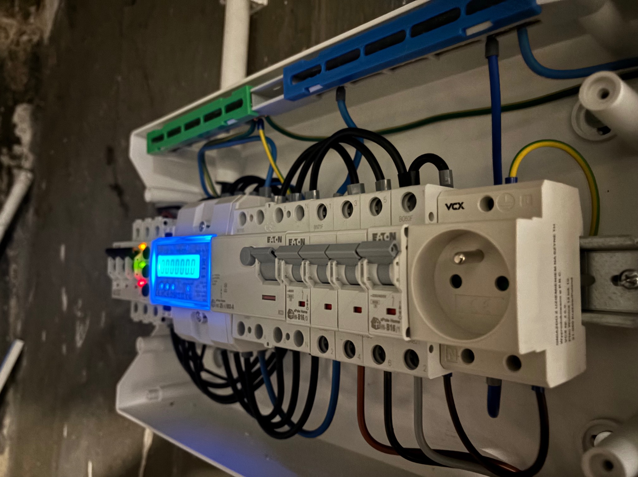 Rozdzielnica elektryczna z licznikiem i gniazdem, widoczne bezpieczniki Eaton, przewody w kolorach: czarnym, niebieskim, żółto-zielonym. Oświetlenie LED licznika.