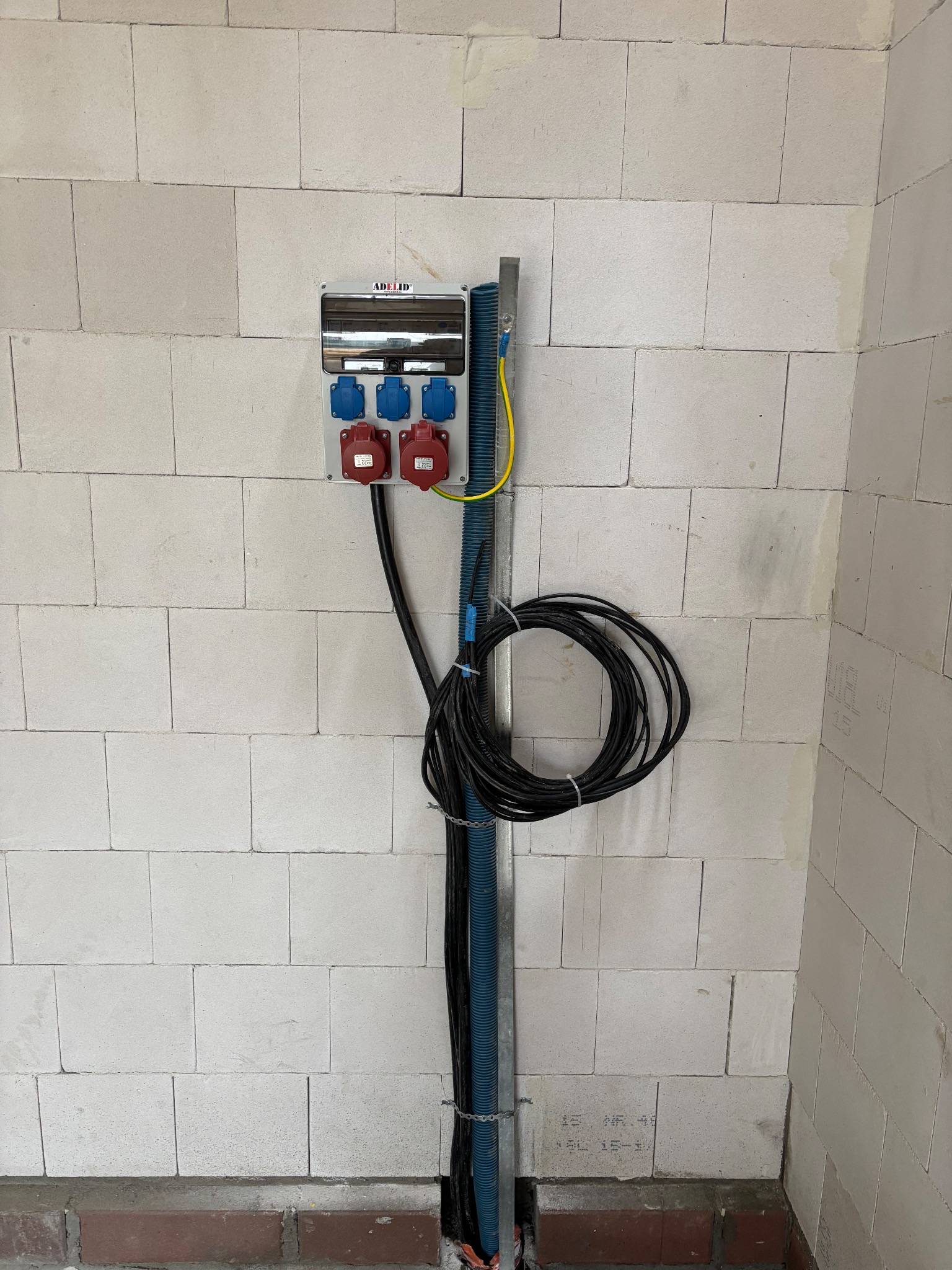 Szara skrzynka elektryczna z gniazdami siłowymi (czerwone i niebieskie) zamontowana na ścianie z białych bloczków. Kable w peszlu prowadzone w dół ściany.