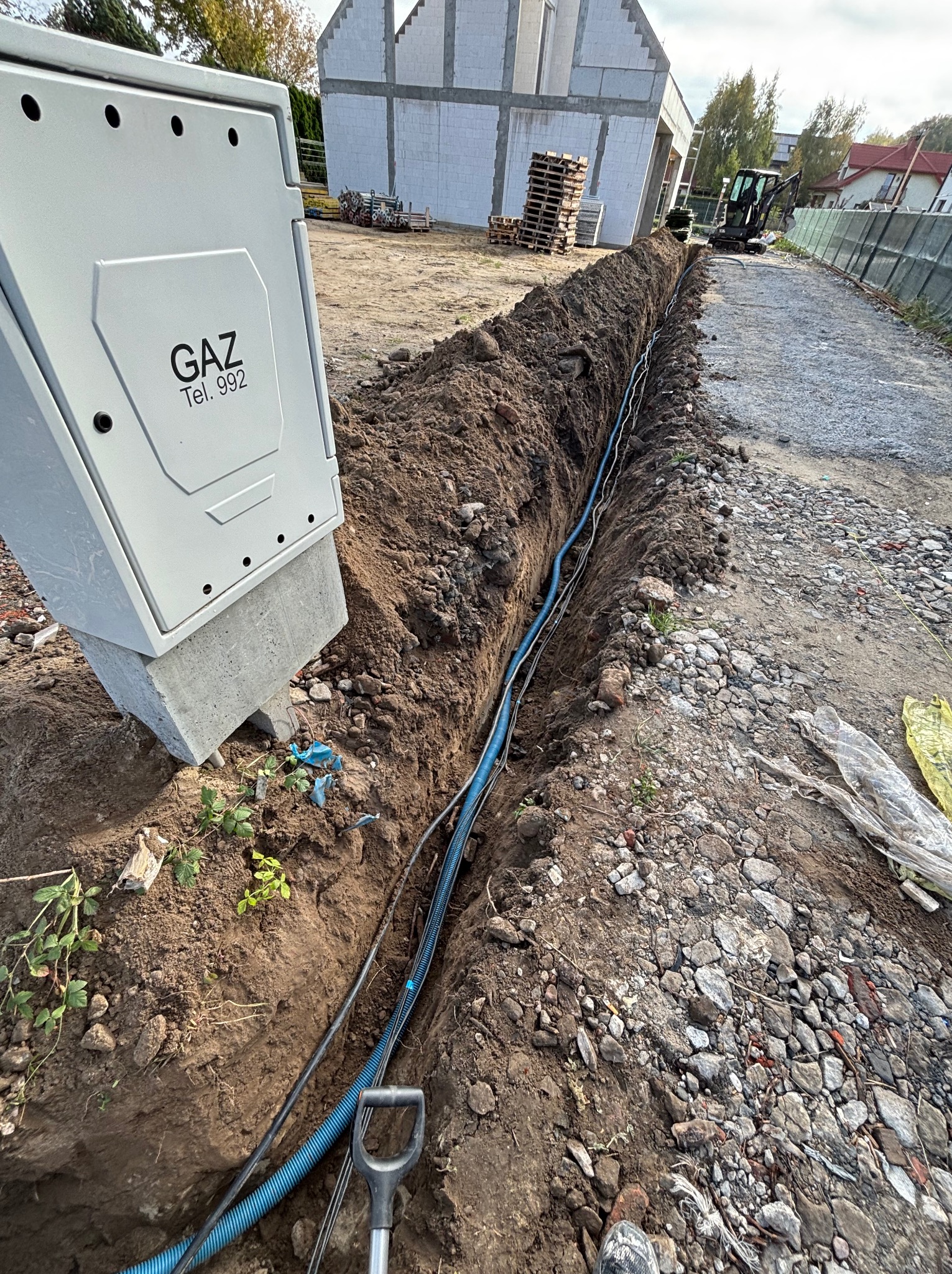 Wykop z ułożonymi kablami (w tym niebieski peszel) w ziemi, obok skrzynka gazowa, w tle budynek w trakcie budowy i koparka. Widok z góry na wykop.