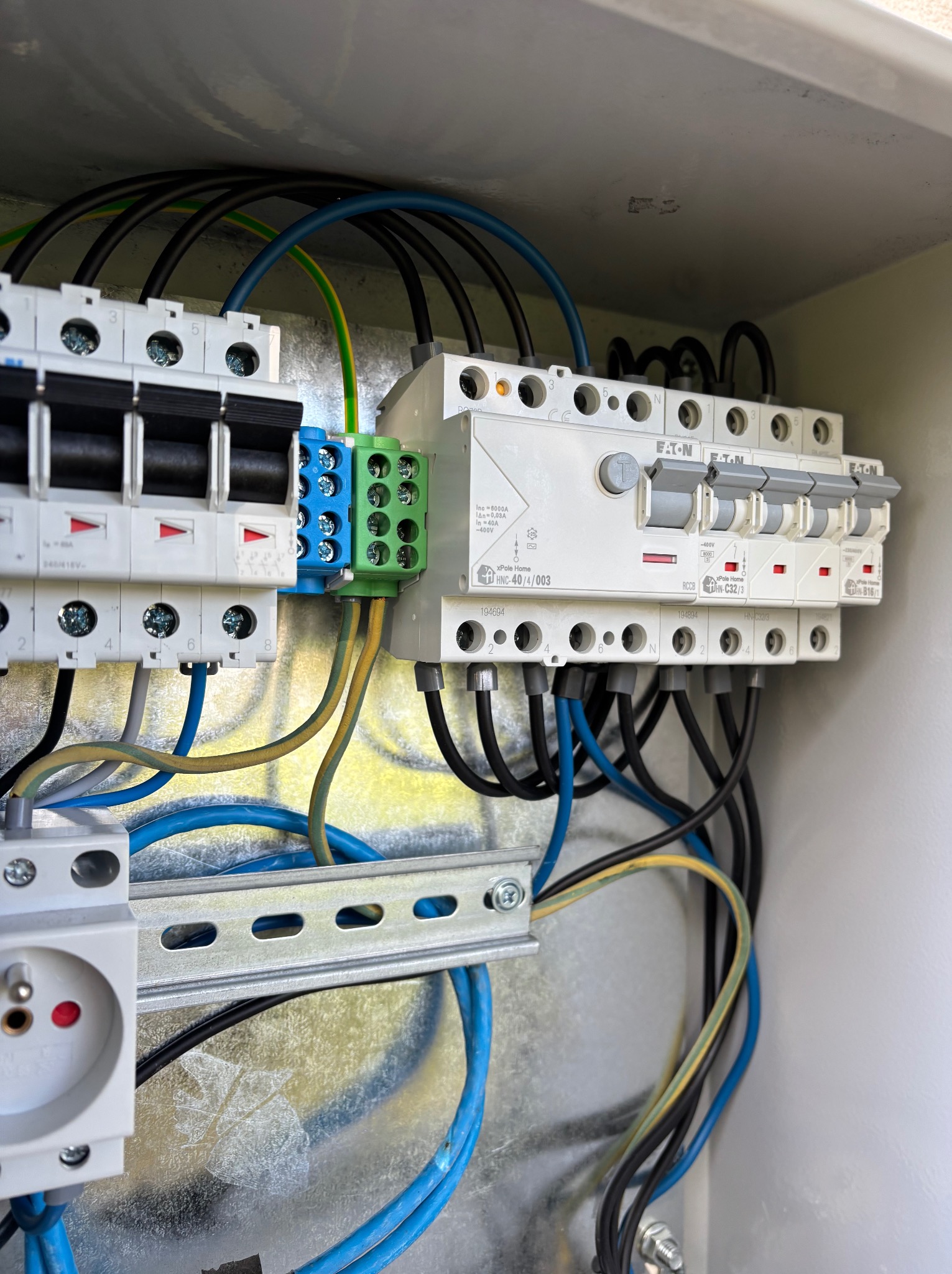 Rozdzielnica elektryczna z bezpiecznikami, przewodami w kolorach: niebieskim, czarnym, żółto-zielonym. Widoczny gniazdko elektryczne i szyna montażowa.