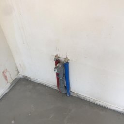 Parit Bud - Wystające ze ściany rury instalacji wodnej (czerwona i niebieska) z szarym trójnikiem, w świeżo wylanym betonowym podłożu, widok z góry.