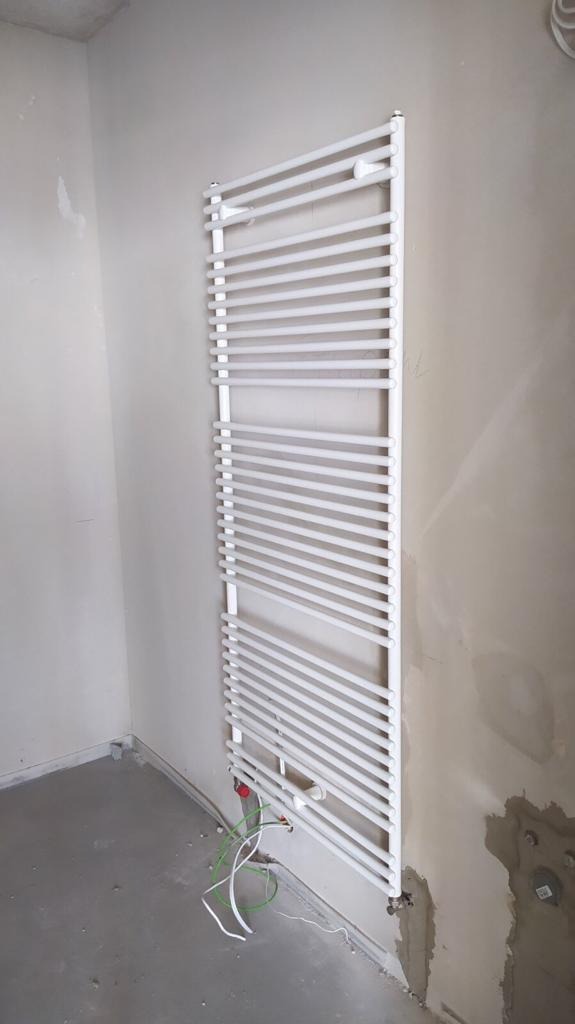 Biały, drabinkowy grzejnik łazienkowy zamontowany na surowej ścianie, widoczne przewody instalacyjne i niedokończone wykończenie podłogi.