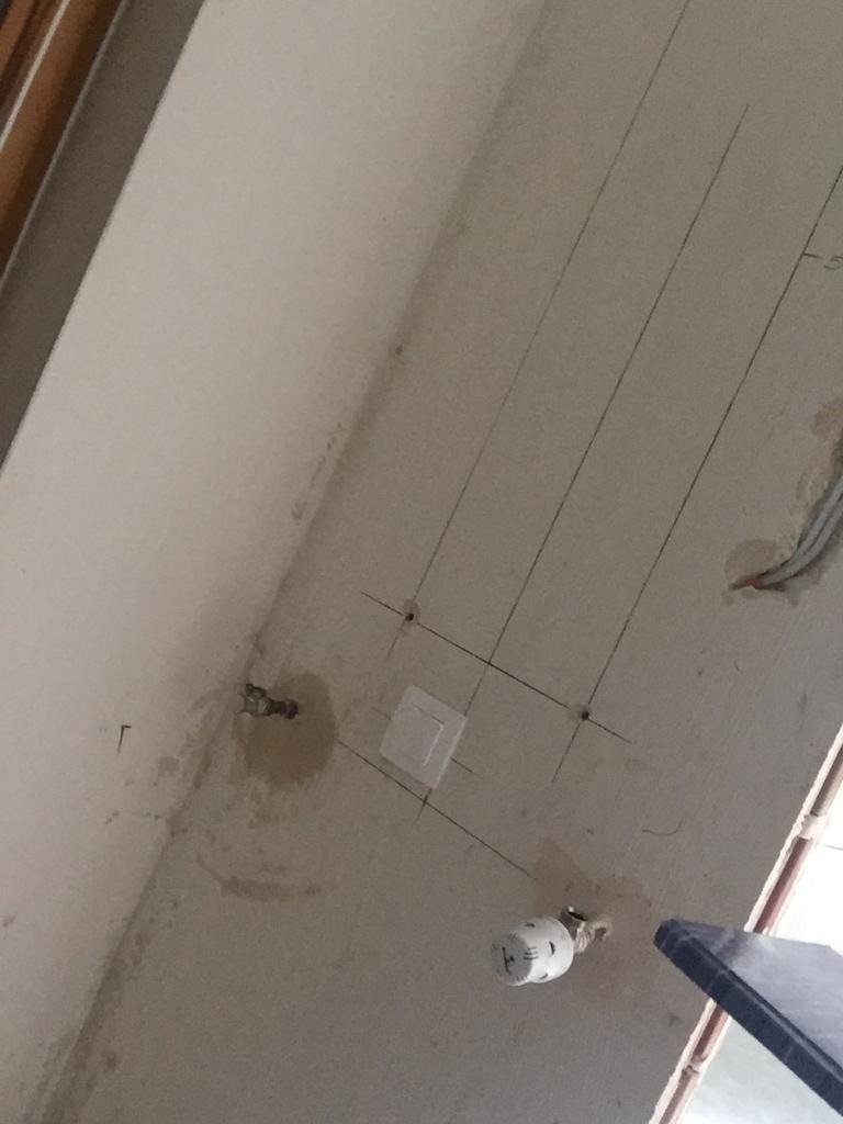 Ściana w trakcie prac hydraulicznych w Warszawie, z widocznymi liniami pomocniczymi, zamontowanym gniazdkiem elektrycznym i wystającymi rurami.