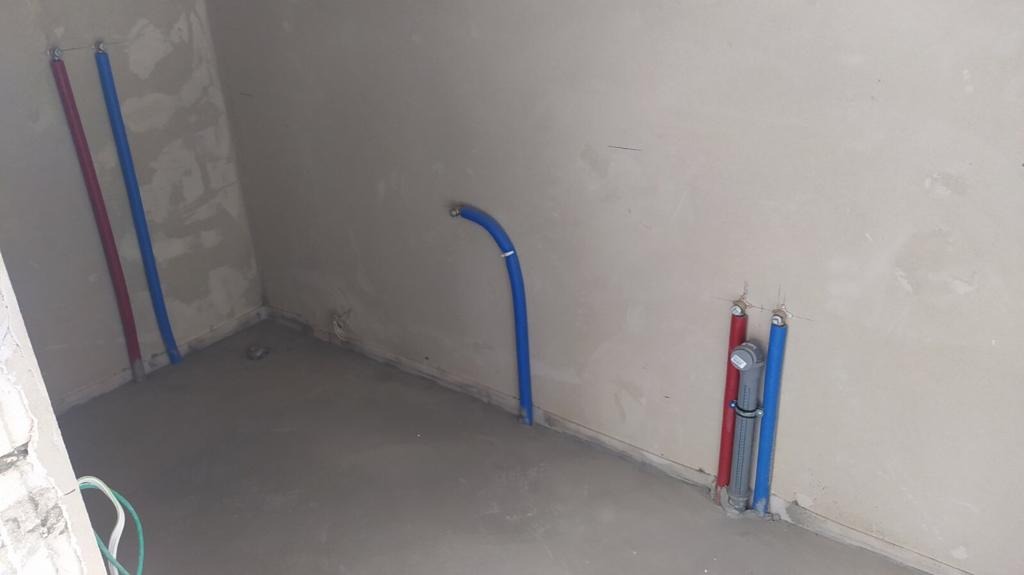 Surowe wnętrze z nową instalacją hydrauliczną: pionowe rury PEX w kolorach czerwonym i niebieskim przy ścianie, dodatkowe rury wystające ze ściany, świeżo wylana wylewka podłogowa.