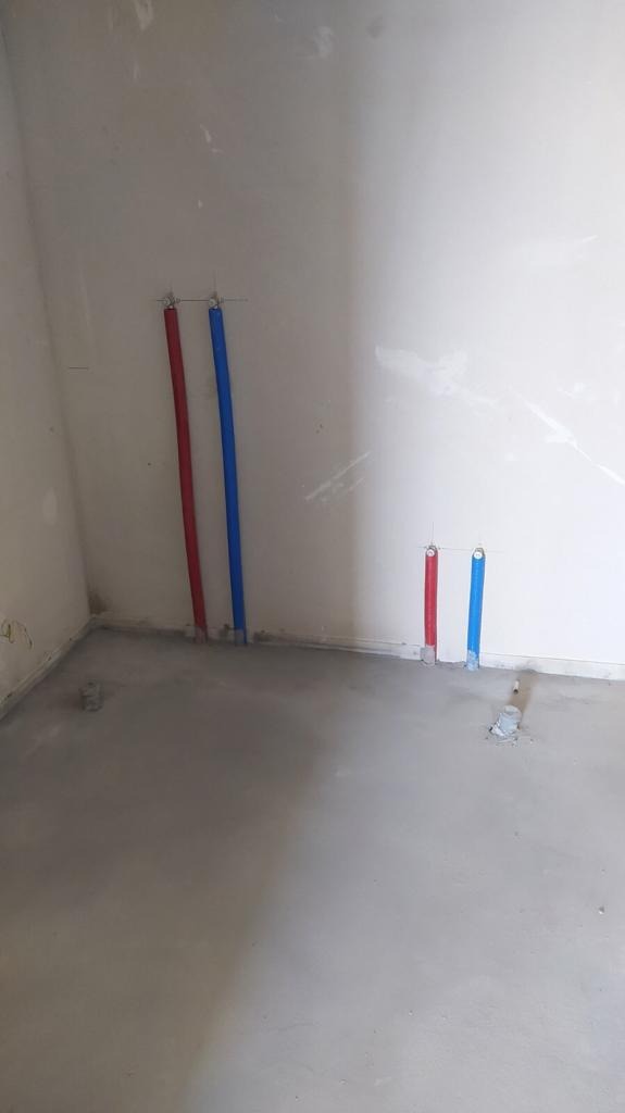Surowy stan łazienki z wylewką betonową i wyprowadzonymi rurami instalacji wodnej w kolorze czerwonym i niebieskim, wystającymi ze ściany.
