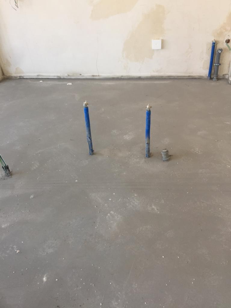Surowy betonowy wylewka podłogowa z wystającymi niebieskimi rurami instalacji wodnej, widoczne ślady po pracach budowlanych na ścianach.