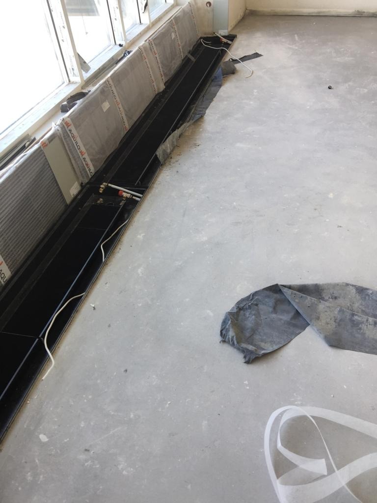 Montaż kanałowych grzejników podokiennych, widoczne rury instalacji grzewczej, folia ochronna na grzejnikach, surowy beton na podłodze.