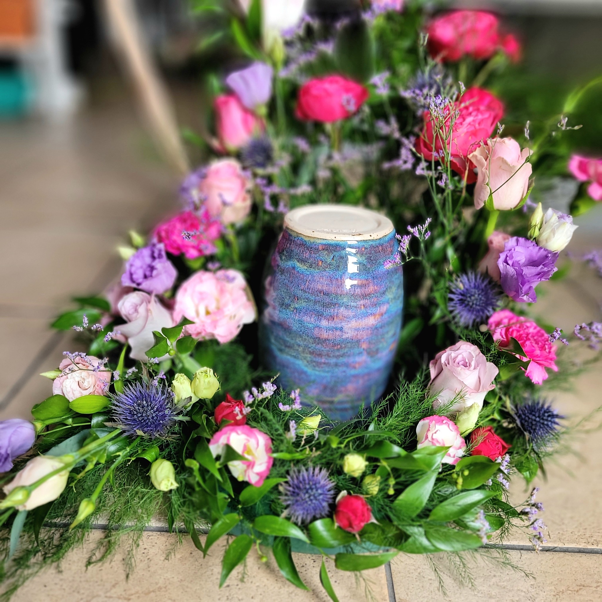 Kompozycja kwiatowa w kształcie wieńca z róż, eustomy i przegorzanu, okalająca ceramiczną urnę w odcieniach błękitu i fioletu, ustawiona na podłodze z płytek.