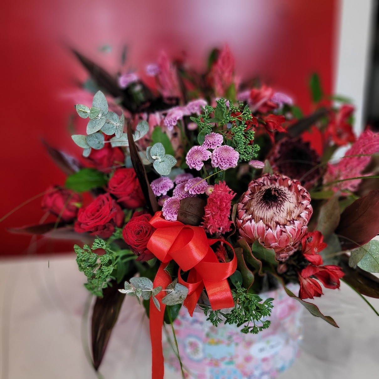 Bogaty bukiet z czerwonych róż, różowych chryzantem i protei królewskiej, przewiązany czerwoną wstążką, umieszczony w ozdobnym pudełku na tle czerwonej ściany.