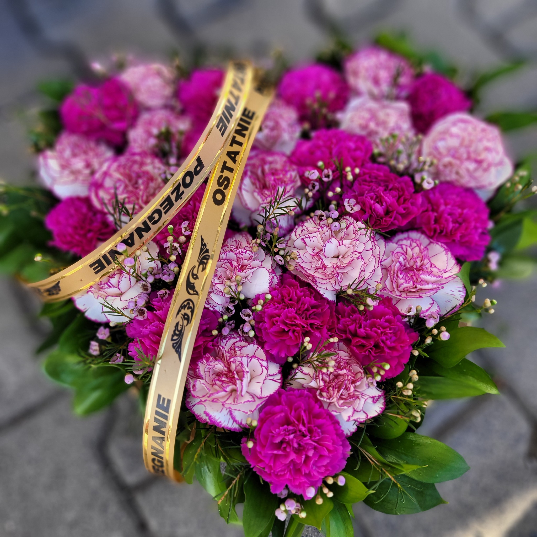 Kompozycja kwiatowa w kształcie serca z różowych i białych goździków, przewiązana złotą wstążką z napisem 'Pożegnanie Ostatnie'.