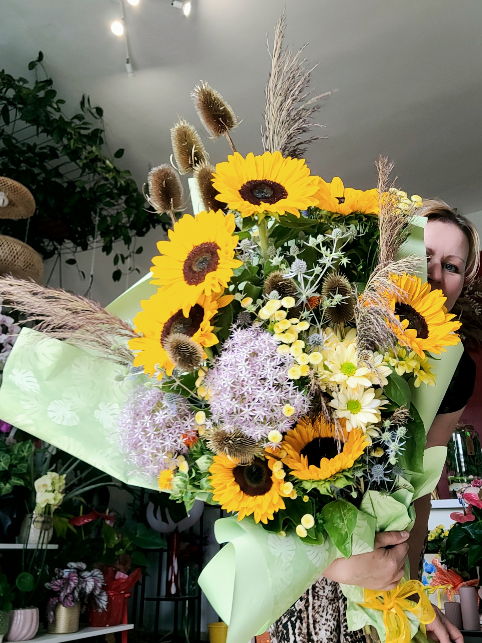 Kobieta trzyma duży, kolorowy bukiet słoneczników, czosnku ozdobnego i suchych traw, owinięty w jasnozielony papier z motywem liści, widoczny w kwiaciarni na tle innych roślin.