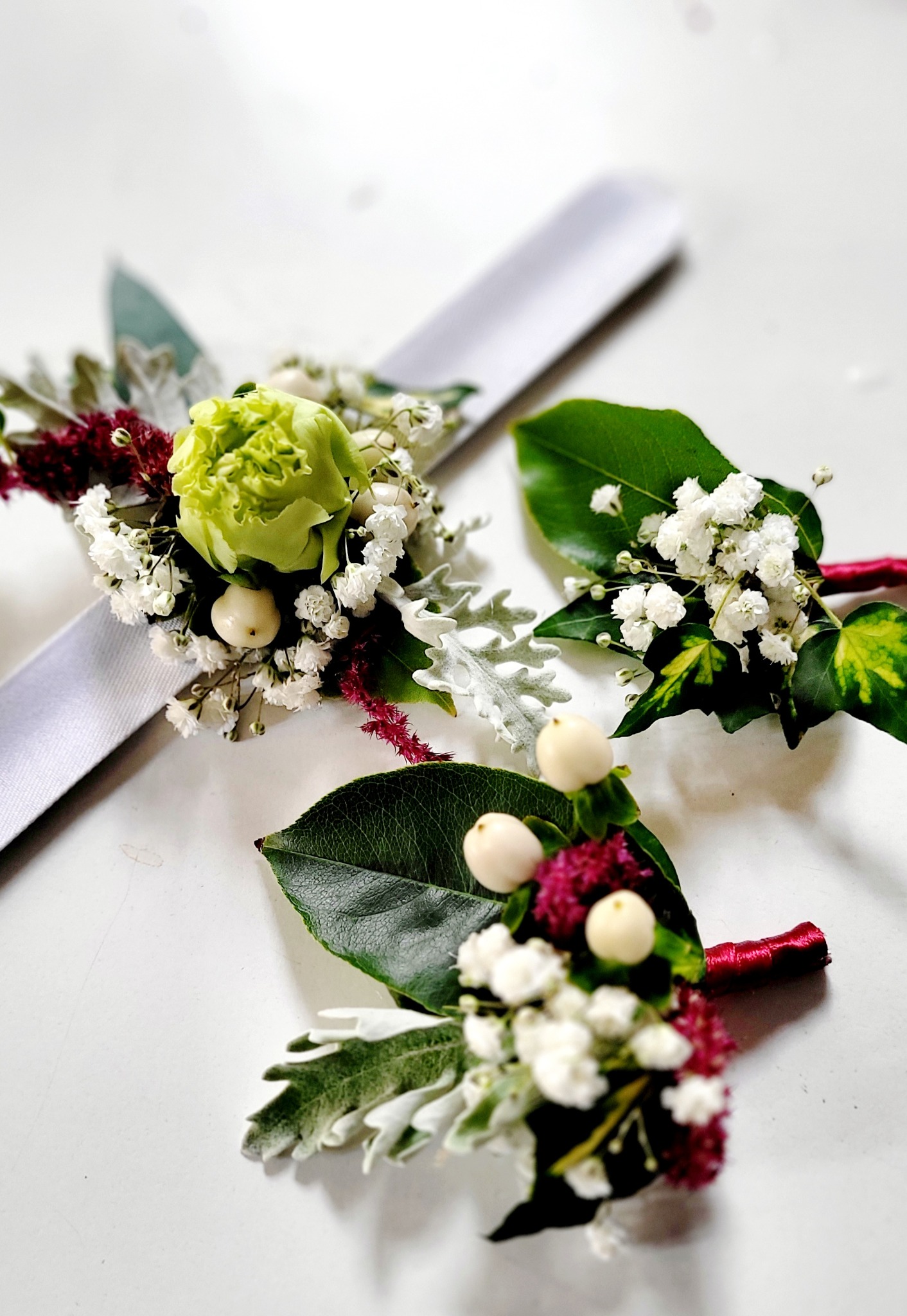 Trzy butonierki ślubne: korsarz na rękę z zieloną różą i białą gipsówką na srebrnej wstążce oraz dwie przypinki do klapy marynarki z białą gipsówką, burgundowymi akcentami i zielonymi liśćmi...