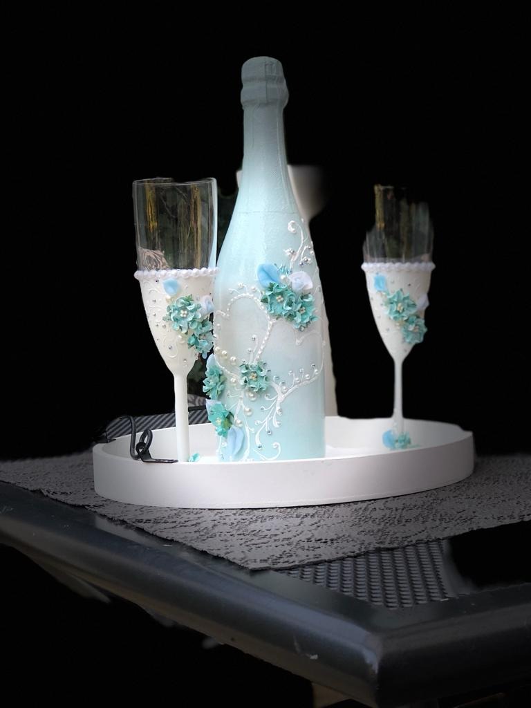 Dekorowana butelka i dwa kieliszki na białej tacy, ozdobione błękitnymi kwiatami i perłami, na ciemnym tle.