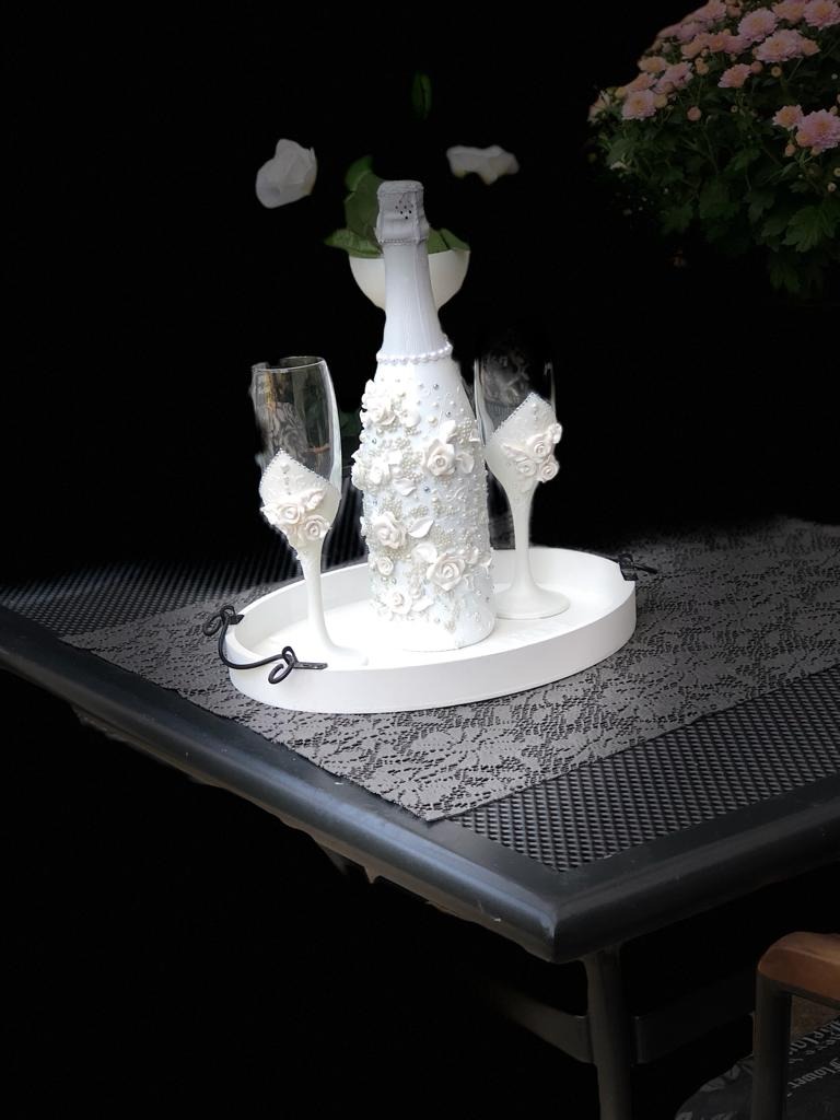 Biała butelka i dwa kieliszki, ozdobione białymi kwiatami i perłami, ustawione na białej tacy z czarnymi uchwytami, na stole z szarą, ażurową serwetą, w tle ciemne tło z roślinami i kwiatami.