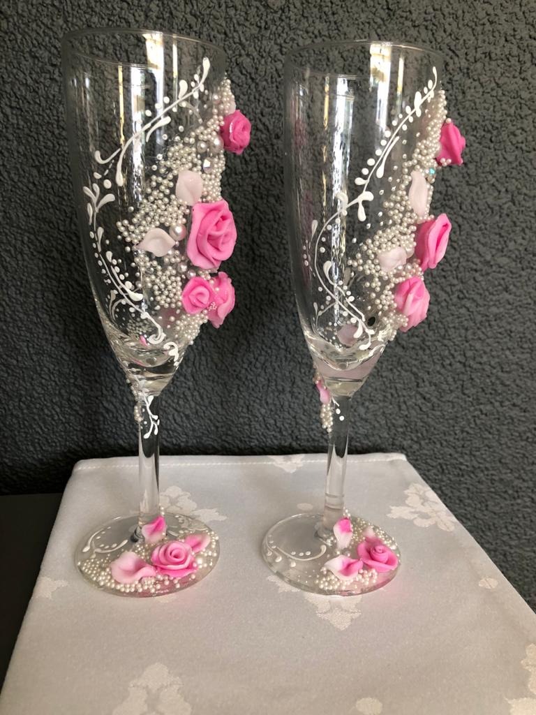 Dwa kieliszki do szampana, ręcznie zdobione białymi ornamentami, perłami i różowymi różyczkami z lukru, stojące na białym obrusie z delikatnym wzorem.