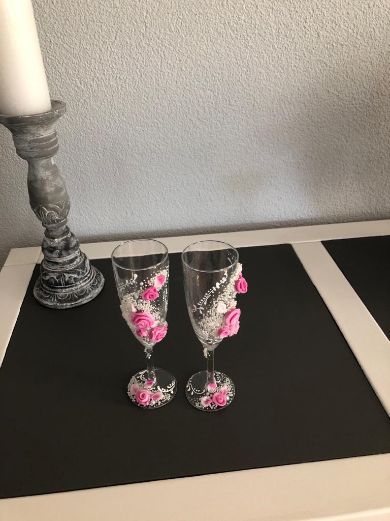 Dwa kieliszki do szampana ozdobione małymi różowymi różami i białymi koralikami, stojące na czarnym blacie obok ozdobnego świecznika.