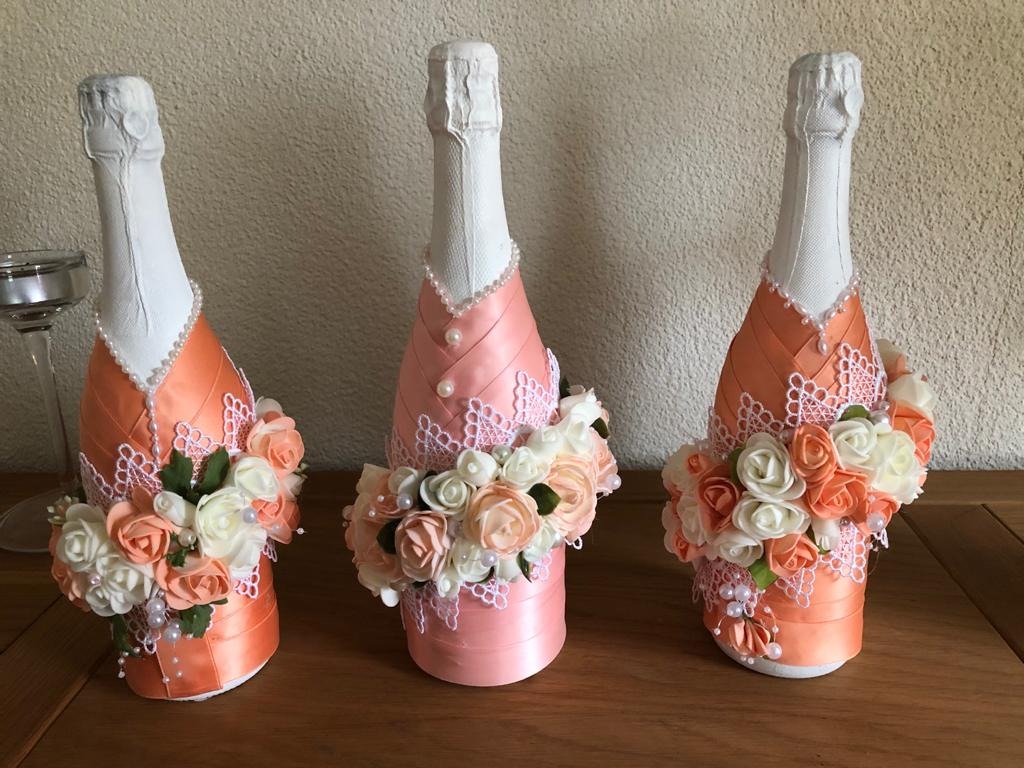 Trzy butelki szampana, każda ozdobiona satynową wstążką w odcieniach brzoskwini i różu, koronką, perłami i bukietem kremowych i brzoskwiniowych róż, stojące na drewnianym stole.