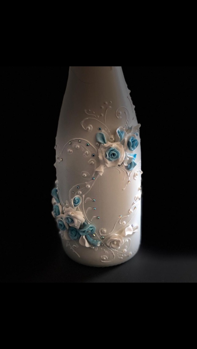 Dekoracyjna, biała butelka z ręcznie robionymi, błękitnymi i białymi różami oraz perłowymi zdobieniami na ciemnym tle.