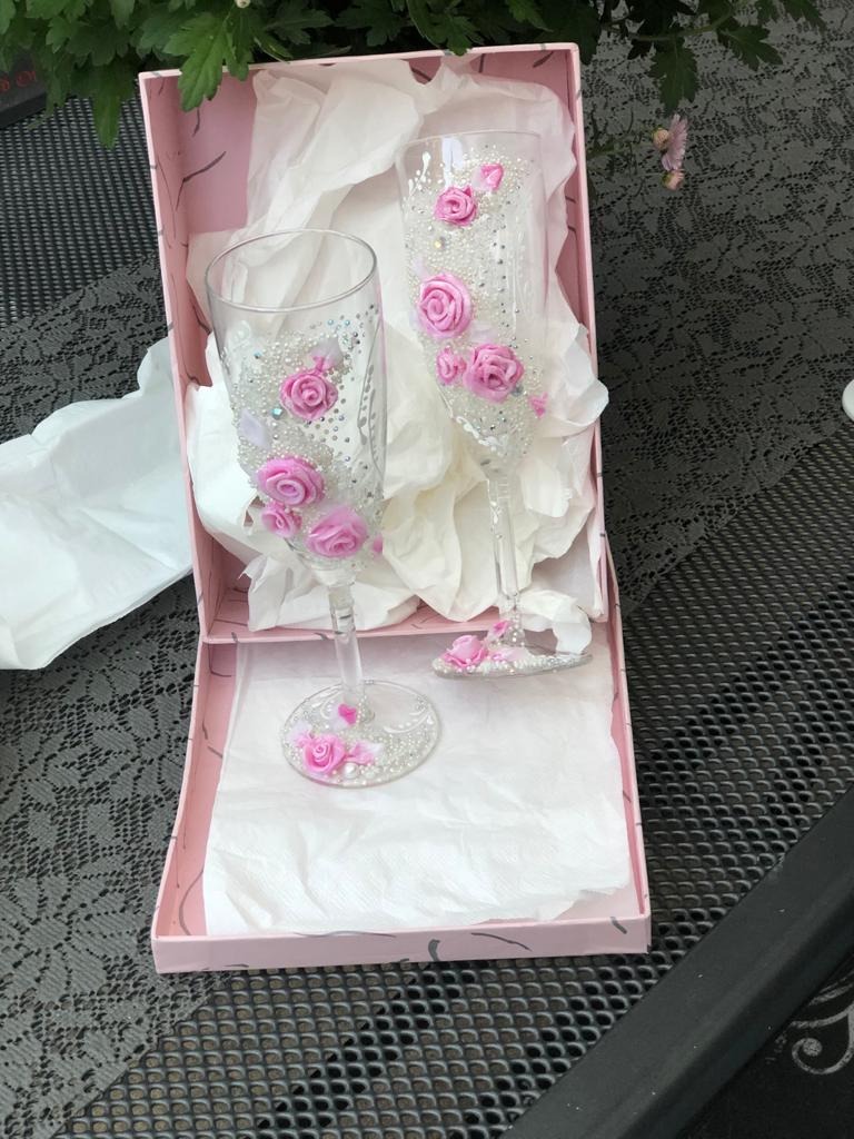 Dwa kieliszki do szampana w ozdobnym, różowym pudełku, udekorowane drobnymi koralikami i różowymi różyczkami, leżące na białym papierze.