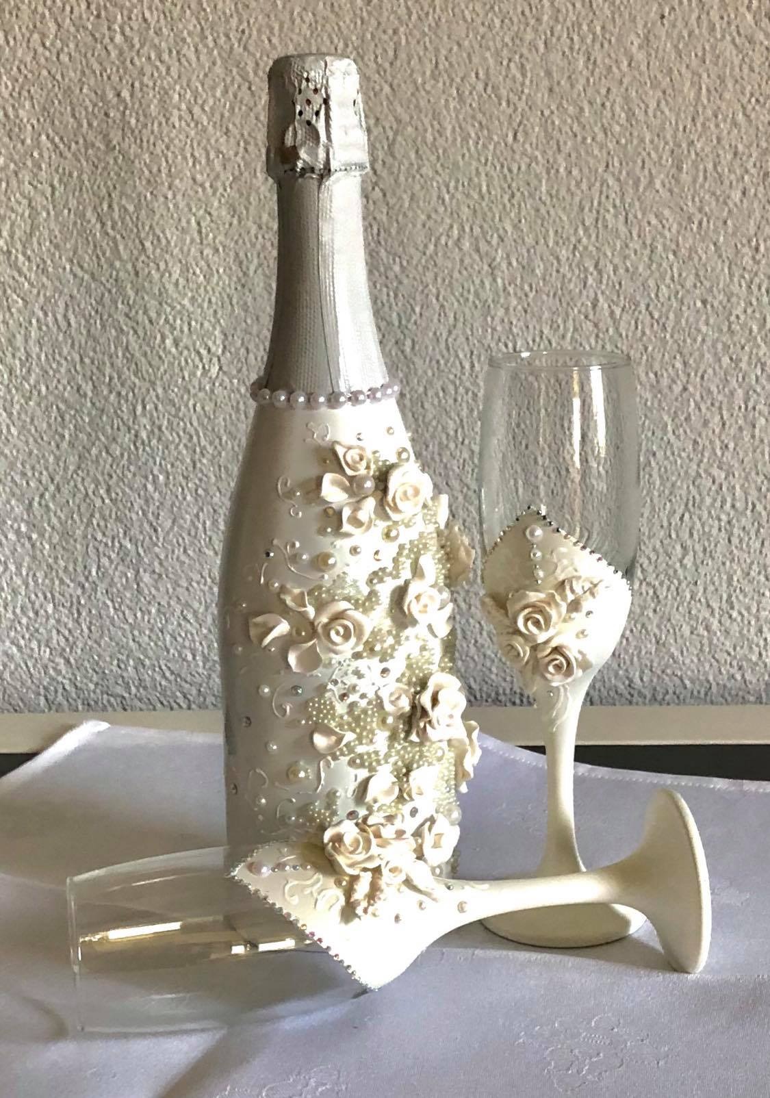 Dekorowana butelka szampana i dwa kieliszki, zdobione kremowymi różami, perłami i drobnymi kryształkami, leżące na białym obrusie.
