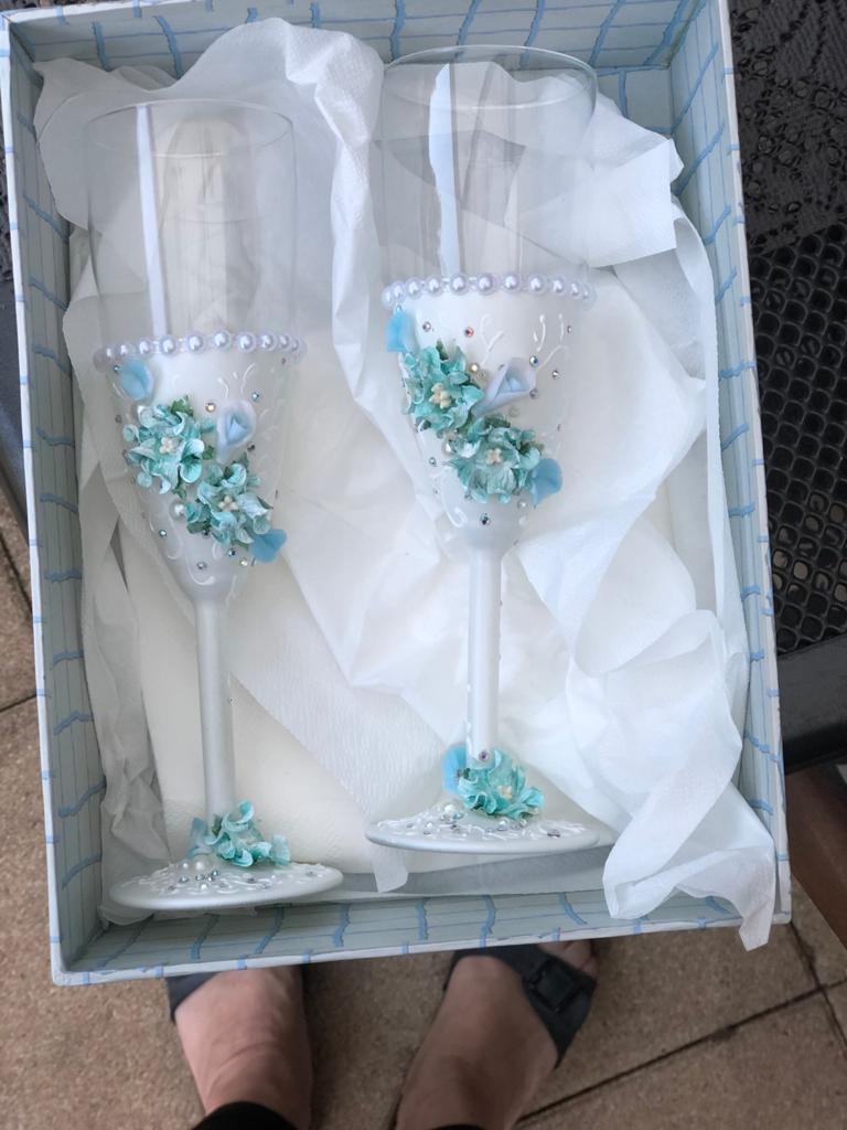 Dwa ręcznie zdobione, białe kieliszki do szampana z turkusowymi kwiatami i perłami, ułożone w pudełku z błękitnym wzorem i białym papierem.