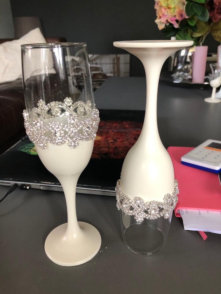 Dwa ozdobne kieliszki do szampana z kryształkami, jeden z białym trzonkiem i podstawą, drugi przezroczysty, ustawione na biurku obok notatnika i kalkulatora.