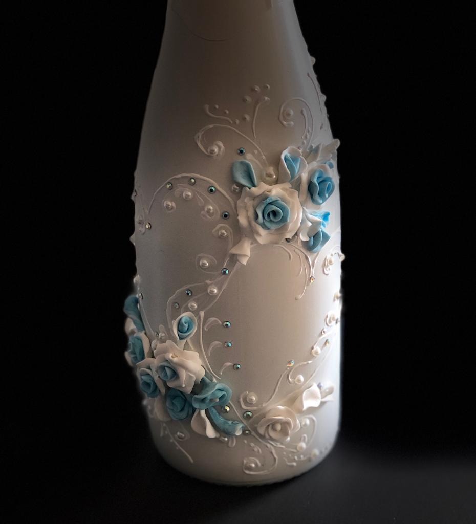 Biała butelka z ręcznie wykonanymi dekoracjami z modeliny: jasnoniebieskie i białe róże, perły i filigranowe ornamenty na czarnym tle.