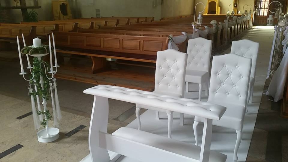 Dekoracja kościoła na uroczystość ślubną: białe krzesła z pikowanymi oparciami i biała ławka, ustawione w rzędzie wzdłuż nawy, dodatkowo świecznik z kompozycją roślinną i świecami.