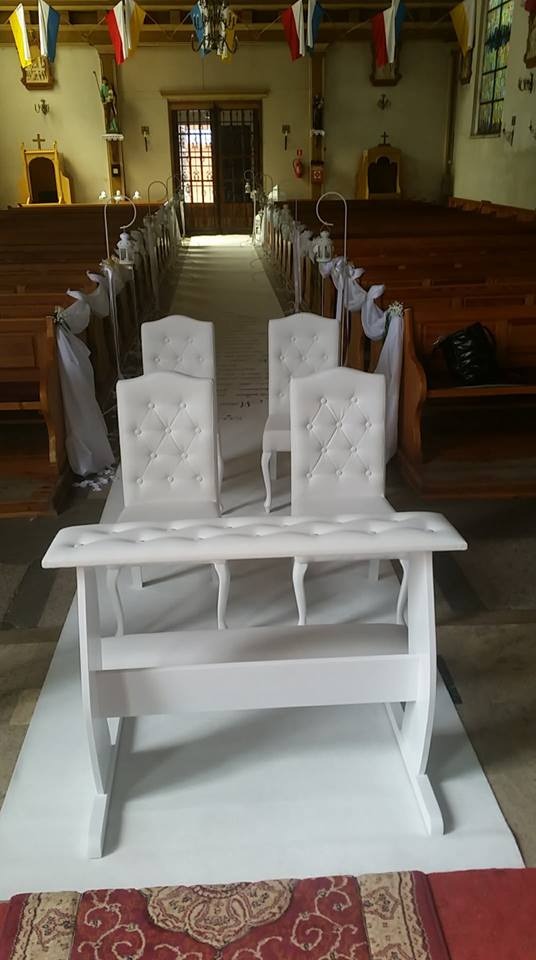 Białe krzesła z pikowanymi oparciami i biała ławka w kościele, przygotowane na uroczystość ślubną, z dekoracjami z białych wstążek na ławkach.