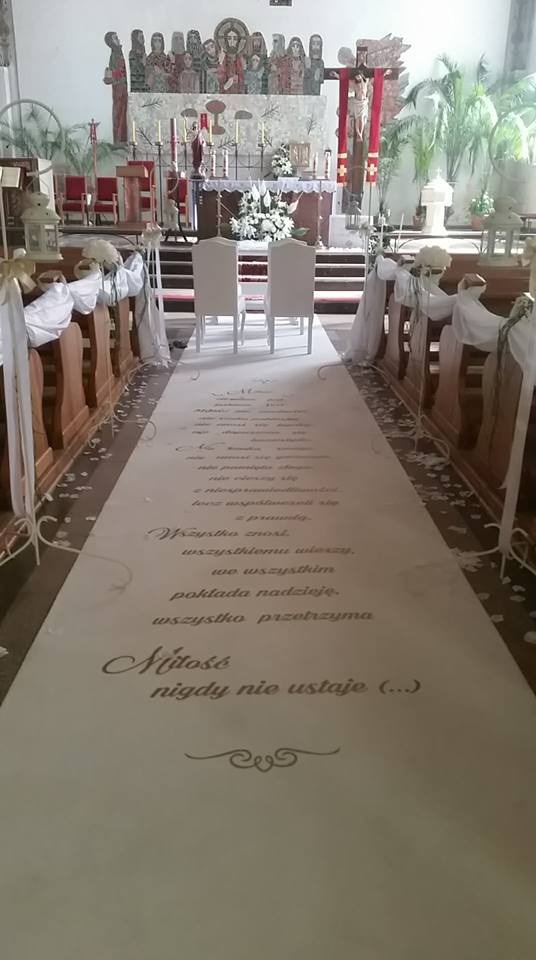 Dekoracja kościoła przed ślubem: biały dywan z cytatem o miłości, białe krzesła i kwiaty na ołtarzu.