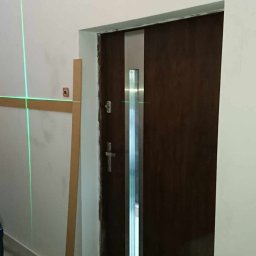 Rafał Wołowczyk - Montaż drzwi z wykorzystaniem poziomicy laserowej, widoczne listwy przypodłogowe i fragment schodów z metalową balustradą.