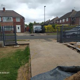 Rafał Wołowczyk - Nowoczesna, pozioma brama wjazdowa z szarych profili, częściowo zamontowana przed domem jednorodzinnym z widocznym samochodem w garażu w tle, teren po budowie, trawnik i żółty kabel na pierwszym...