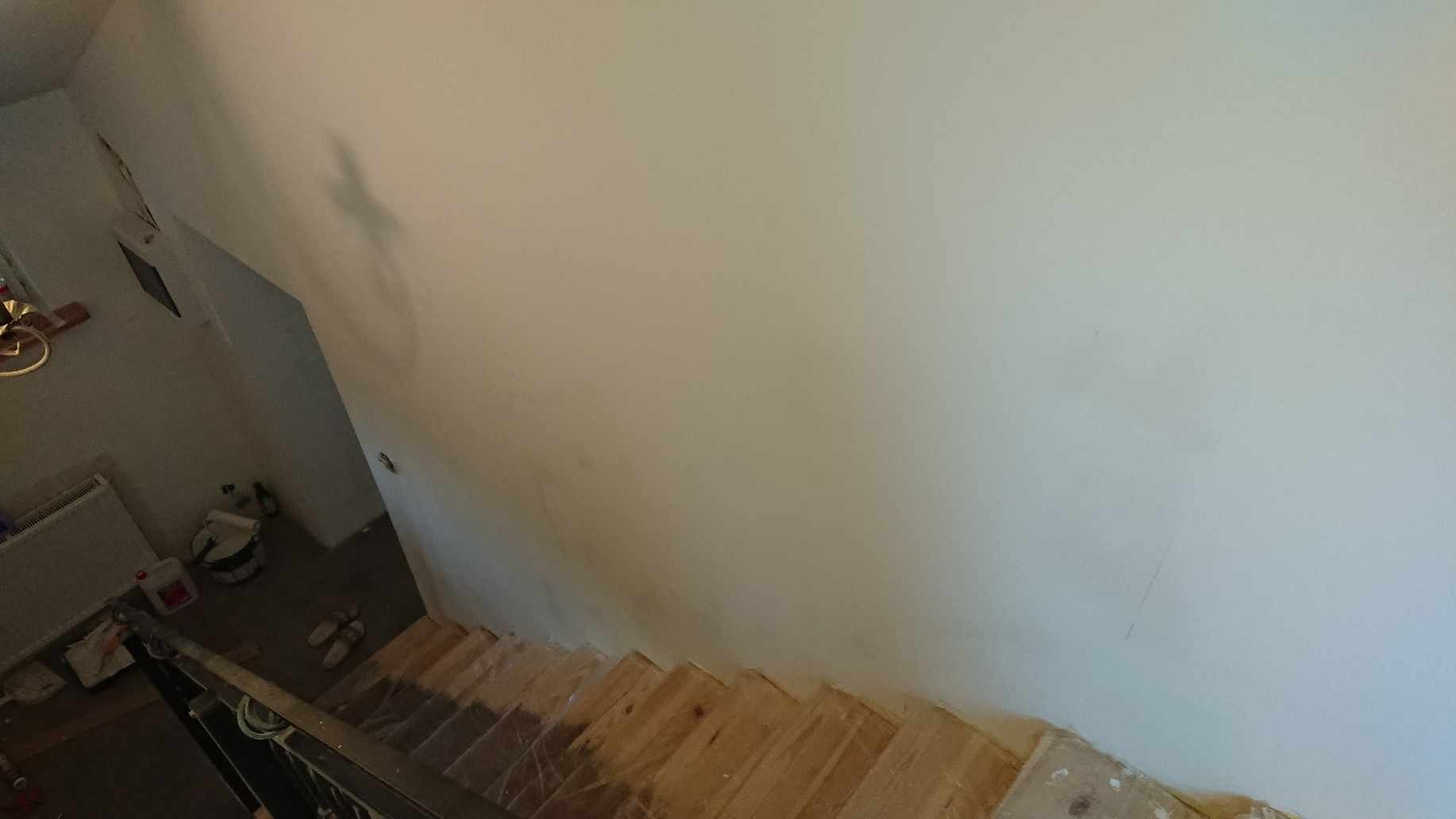 Widok z góry na klatkę schodową w trakcie prac wykończeniowych, z surowymi drewnianymi stopniami i świeżo pomalowaną ścianą w jasnym odcieniu, widoczne ślady po malowaniu na stopniach, w tle wiadro...
