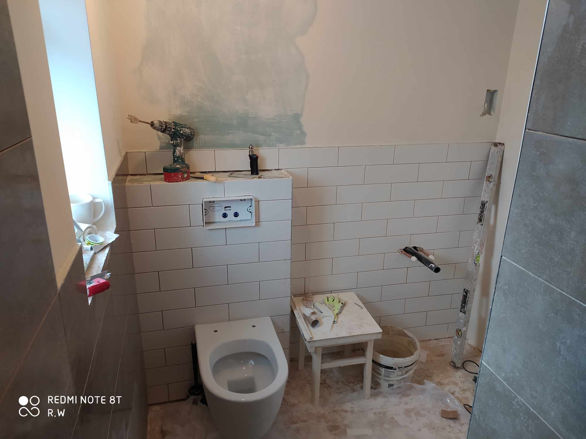 Wnętrze łazienki w trakcie prac wykończeniowych: białe płytki ułożone na ścianie, zamontowany stelaż podtynkowy WC, widoczny otwór na rurę, poziomica oparta o ścianę, narzędzia na taborecie, ślady...