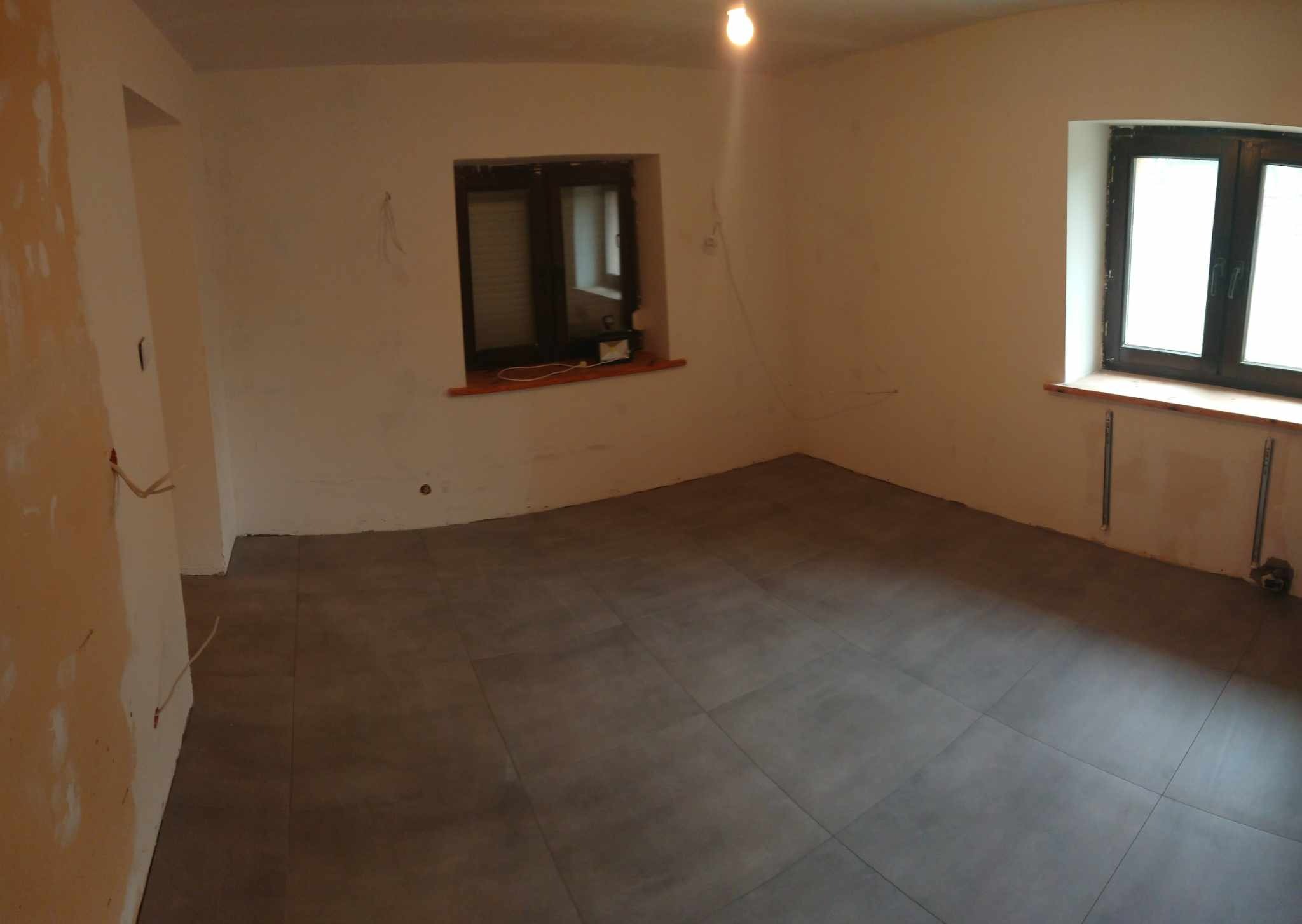 Pokój po ułożeniu szarych płytek podłogowych, ściany w trakcie przygotowania do malowania, widoczne wystające kable i niedokończone wnęki okienne.