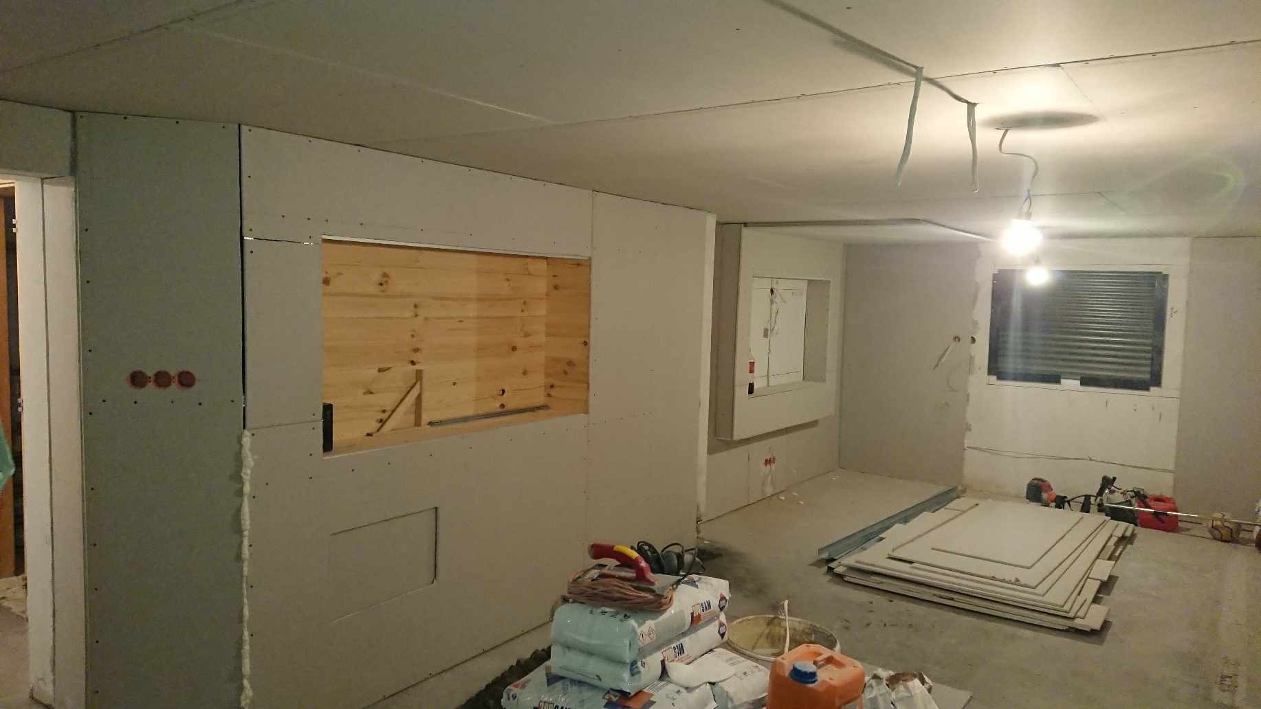 Wnętrze w trakcie prac wykończeniowych: ściany z płyt karton gips, drewniana wnęka w ścianie, widoczne worki z zaprawą i narzędzia.