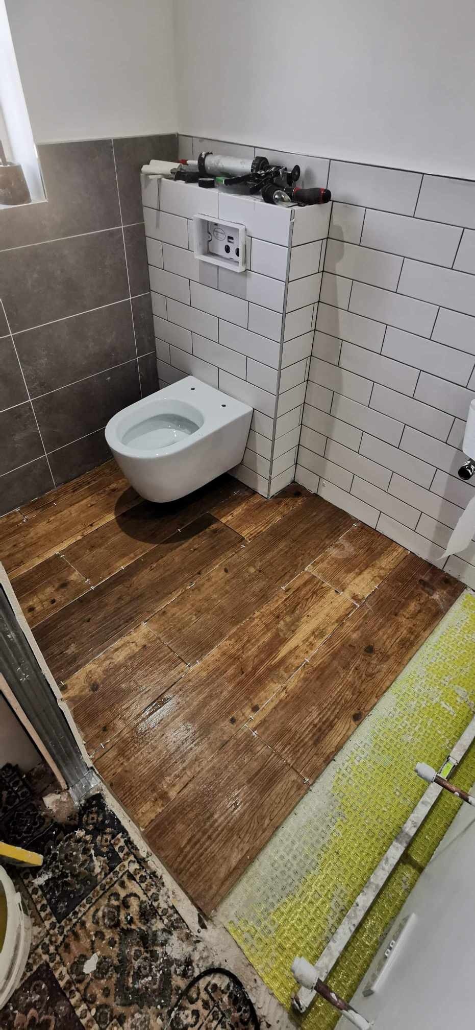 Wnętrze łazienki w trakcie prac wykończeniowych: podwieszana toaleta, drewnopodobne płytki podłogowe z krzyżykami dystansowymi, białe płytki ścienne typu metro, narzędzia na obudowie stelaża.