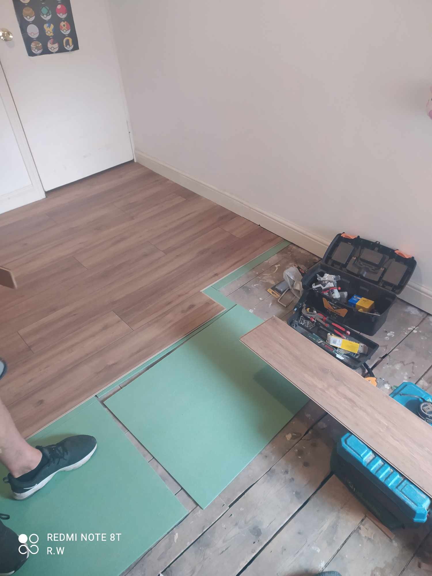 Układanie paneli podłogowych: widoczny fragment ułożonej podłogi, zielone płyty podkładowe, skrzynka z narzędziami, oraz noga w sportowym bucie na tle surowej podłogi.