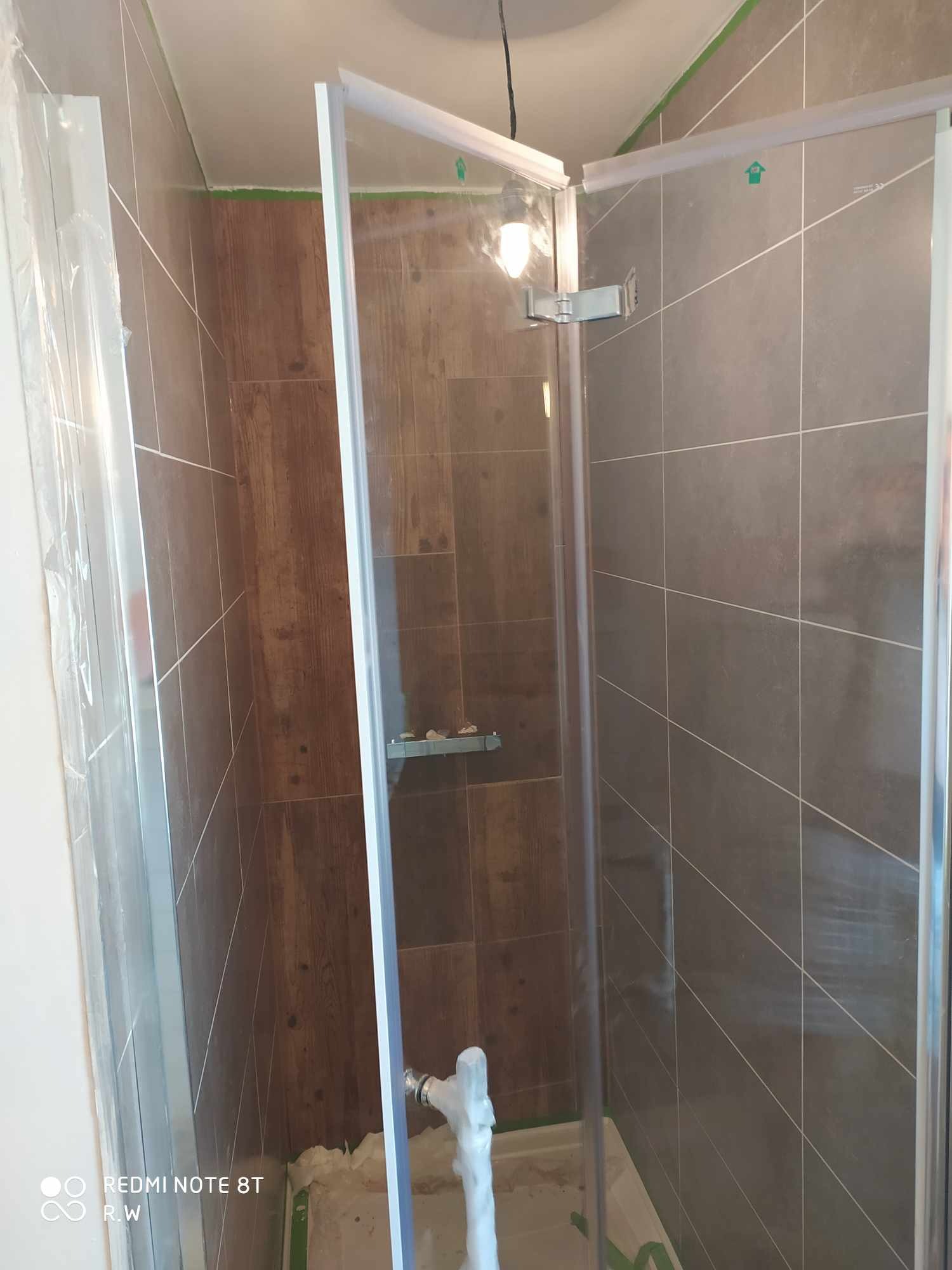 Wnętrze łazienki w trakcie montażu kabiny prysznicowej z widocznym zabezpieczeniem folią i taśmą malarską na elementach oraz fragmentami płytek imitujących drewno i szarych płytek.