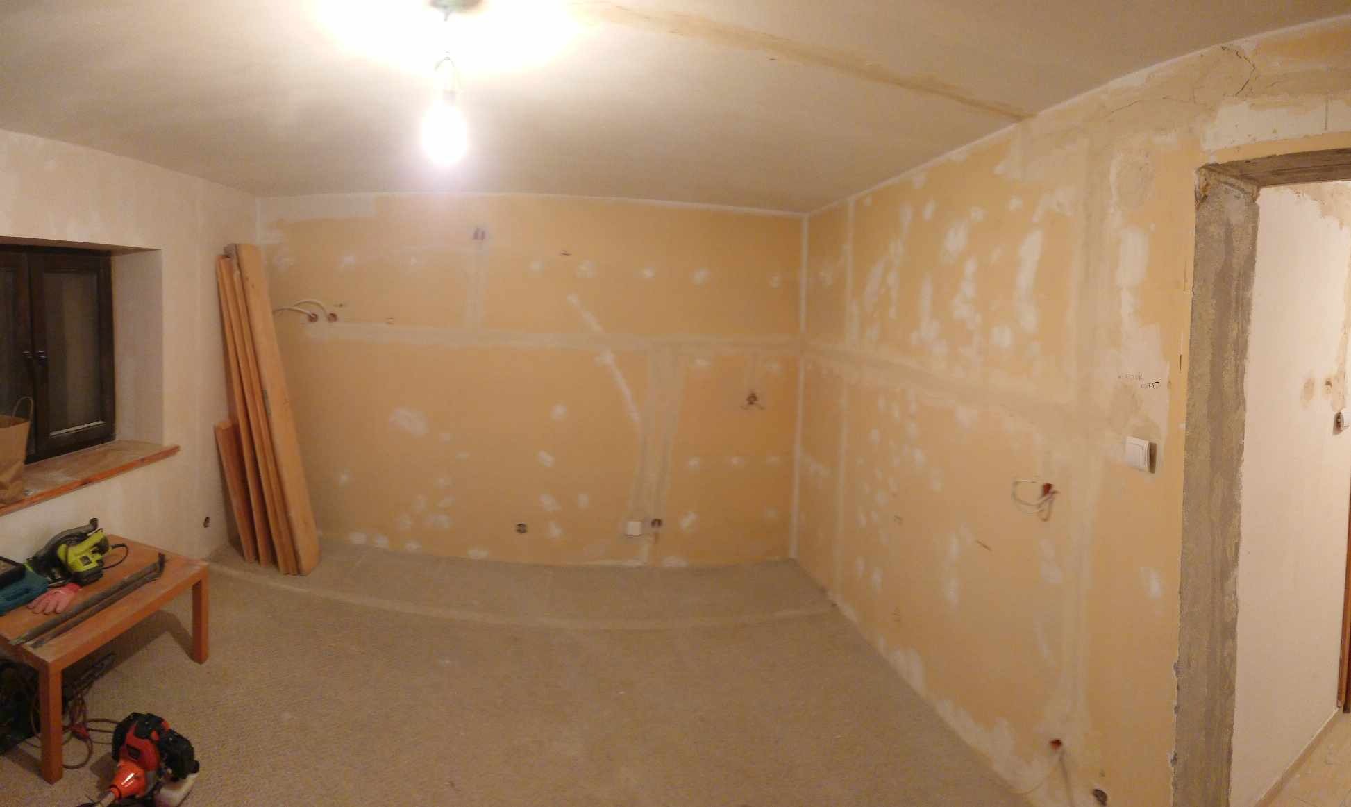 Pokój w trakcie przygotowania do prac wykończeniowych: ściany pokryte gładzią gipsową z widocznymi punktami elektrycznymi, podłoga przygotowana, narzędzia na niskim stoliku przy oknie, deski...