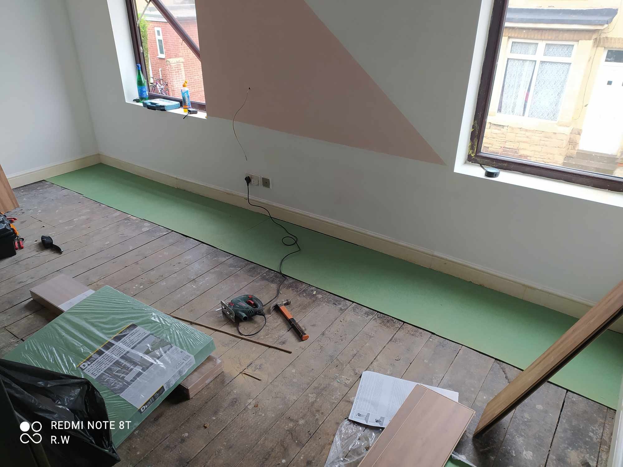 Przygotowanie do układania paneli: zielona warstwa izolacyjna rozłożona na starej, drewnianej podłodze, w tle ściana z geometrycznym wzorem i dwa okna, obok leżą narzędzia - wyrzynarka i młotek.