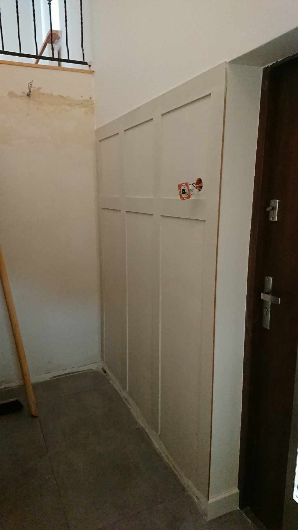 Białe panele ścienne z dekoracyjnymi listwami w przedpokoju, widoczne niedokończone prace przy gniazdku elektrycznym i stara ściana w tle.