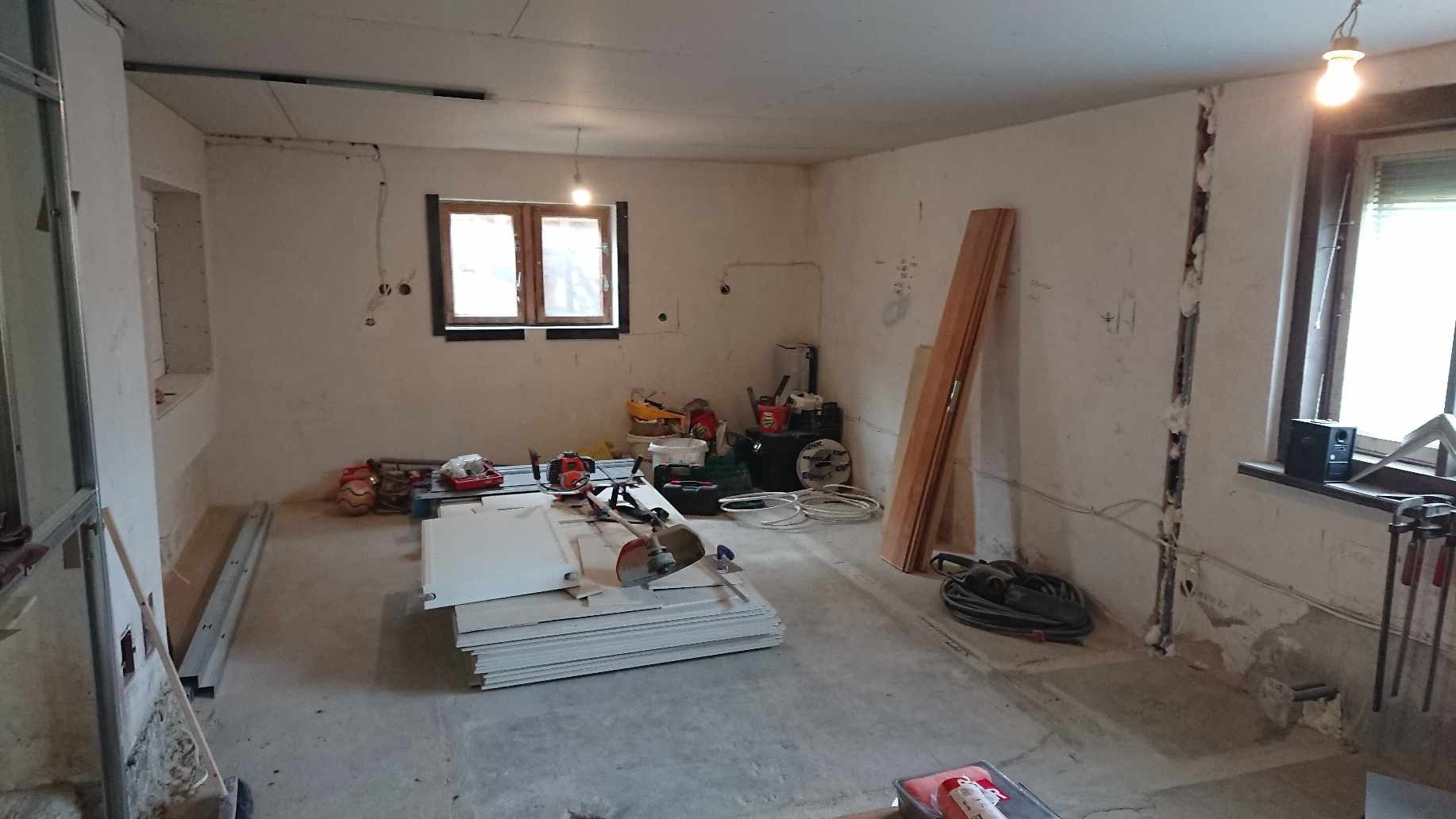 Wnętrze pomieszczenia w trakcie prac przygotowawczych do remontu: gołe ściany, widoczne instalacje, narzędzia, płyty kartonowo-gipsowe i listwy przypodłogowe zgromadzone na podłodze.