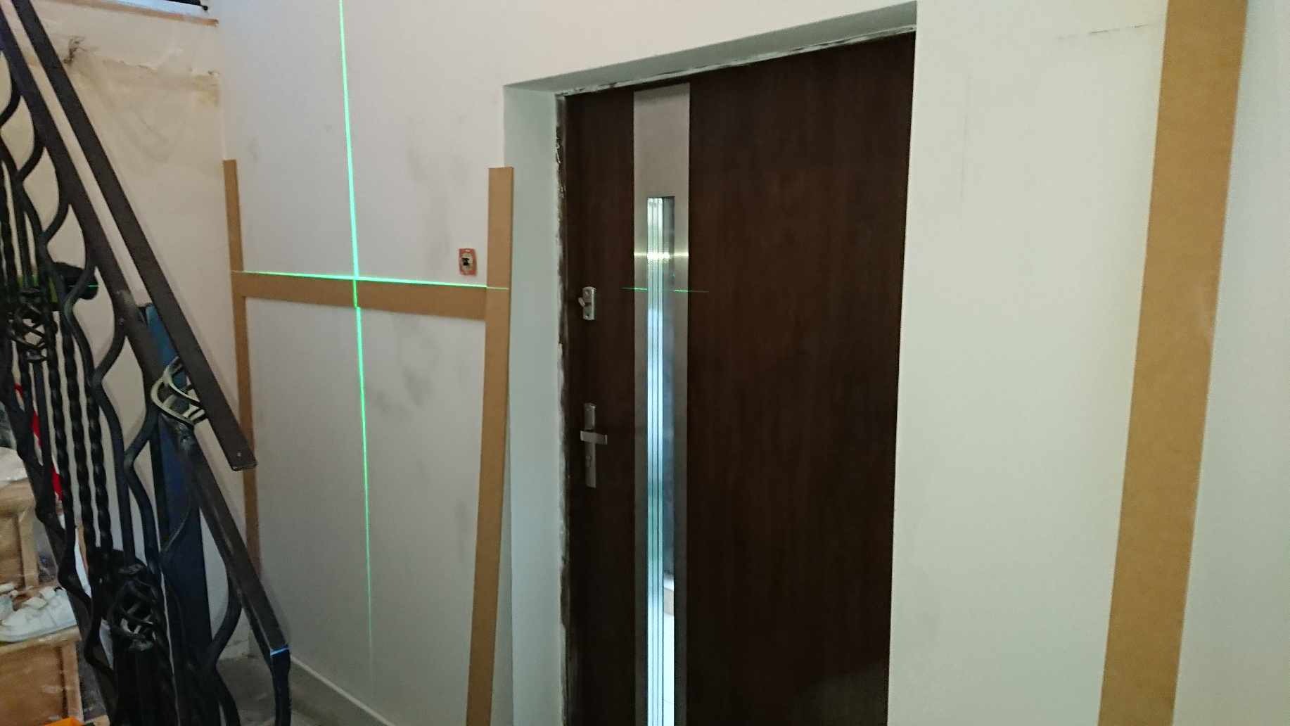 Montaż drzwi z wykorzystaniem poziomicy laserowej, widoczne listwy przypodłogowe i fragment schodów z metalową balustradą.