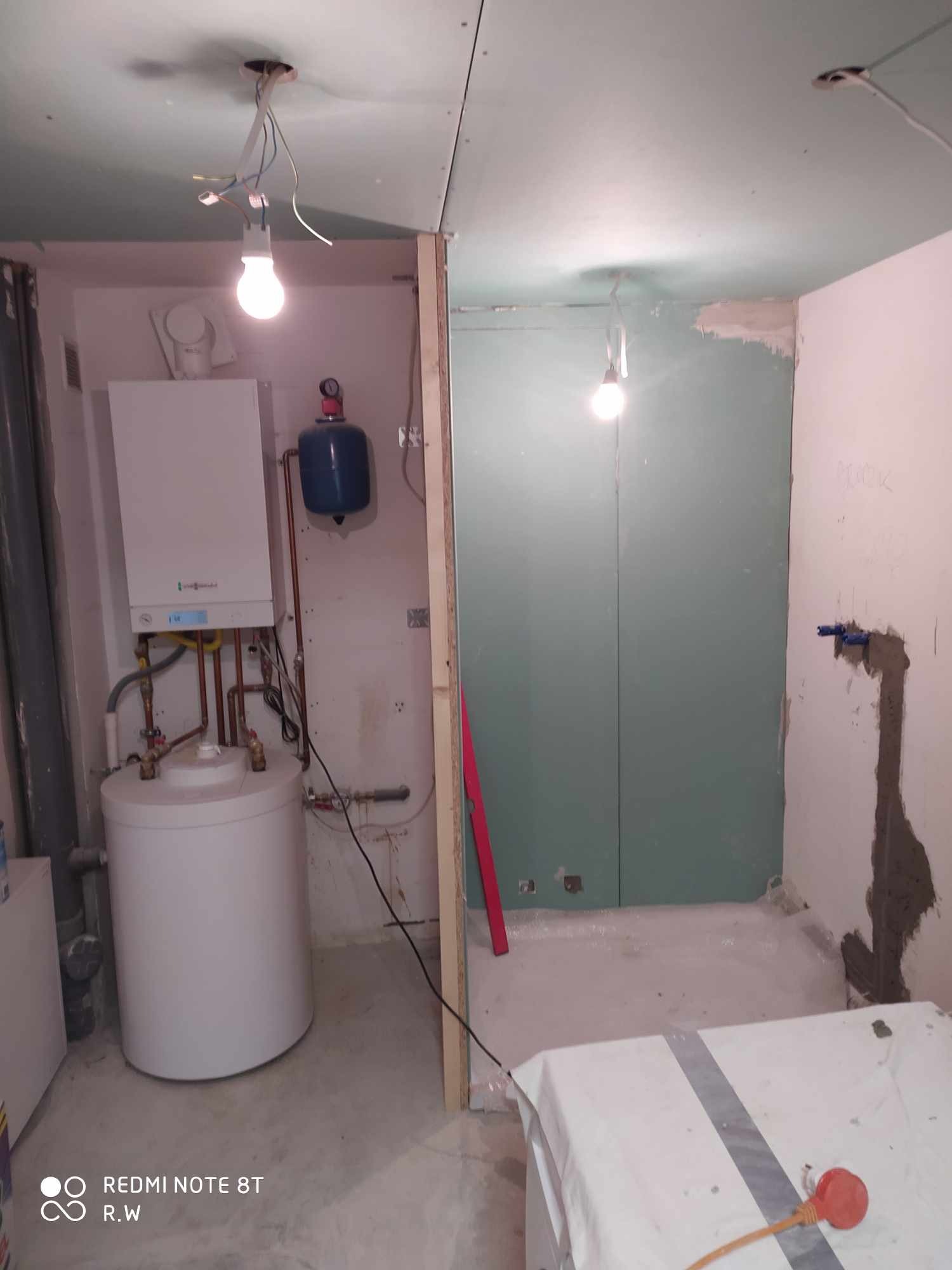 Wnętrze w trakcie prac wykończeniowych: widoczny piec dwufunkcyjny, bojler, częściowo położona płyta karton gips na ścianach, surowy beton na podłodze, wiszące żarówki.