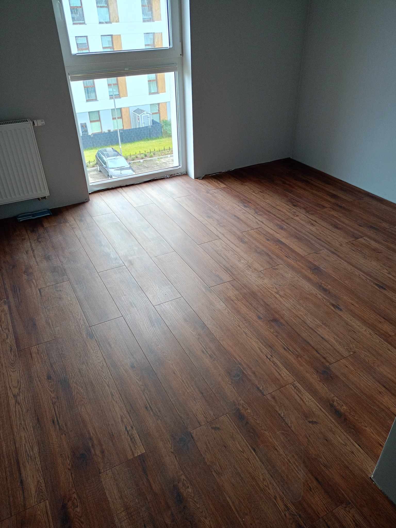 Ułożone panele podłogowe w nowym mieszkaniu, widok z góry na brązowe panele z wyraźną strukturą drewna, ściany pomalowane na szaro, okno z widokiem na parking i budynek.