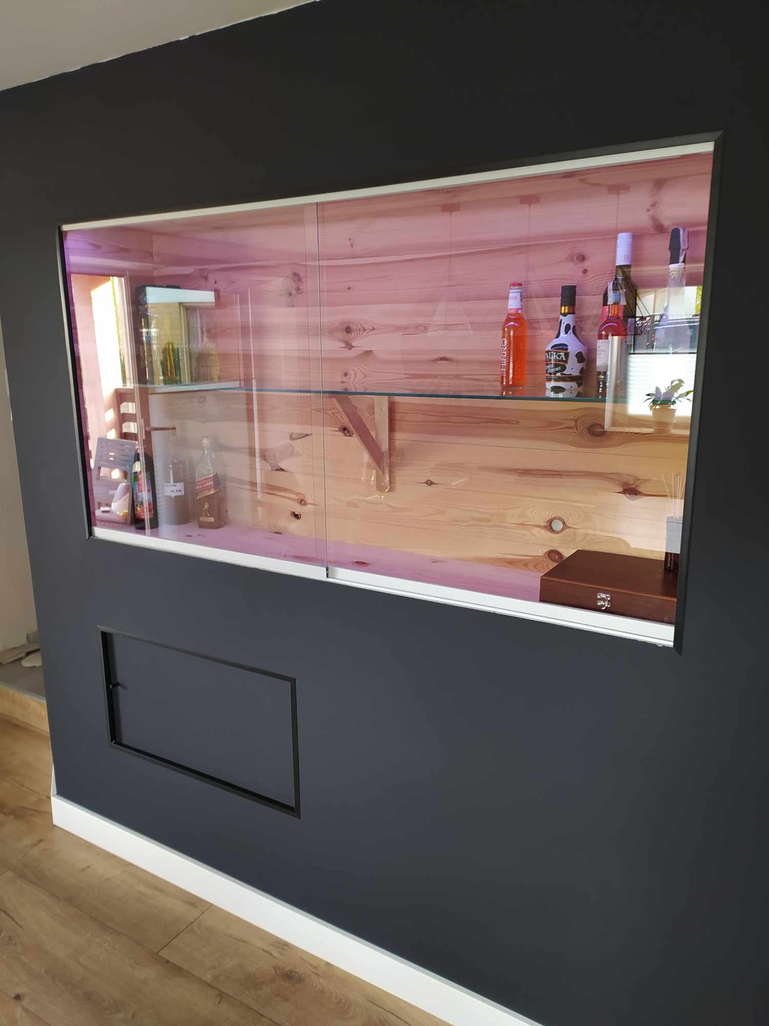 Wnęka ścienna z drewnianym wnętrzem, przeszklonymi przesuwnymi drzwiami, podświetlona, z ekspozycją butelek alkoholi i ozdobnym pudełkiem, na tle szarej ściany z listwą przypodłogową.