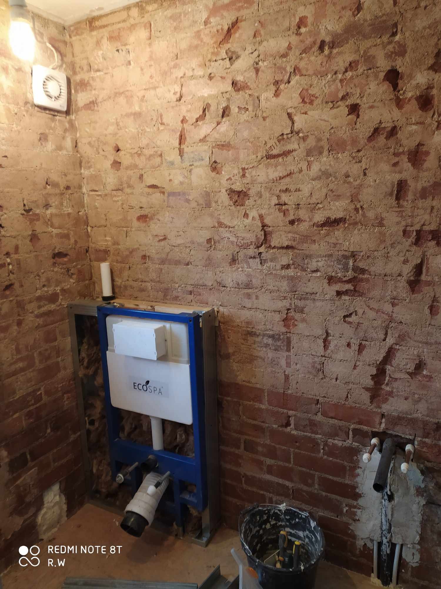 Niezatynkowana ściana z czerwonej cegły w łazience, z widoczną instalacją podtynkową WC w niebieskiej ramie i białą spłuczką EcosPA oraz fragmentem instalacji wodnej i kanalizacyjnej.