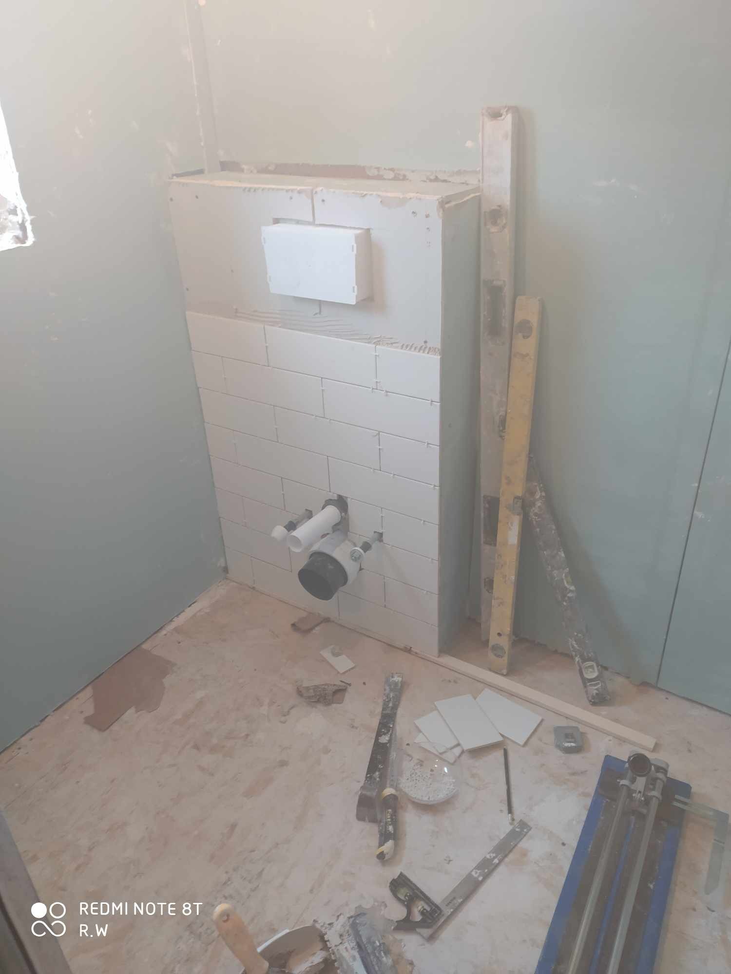 Montaż białych, prostokątnych płytek w łazience na zabudowie stelaża WC, widoczne rury kanalizacyjne, poziomice i narzędzia glazurnicze na podłodze w trakcie pracy.