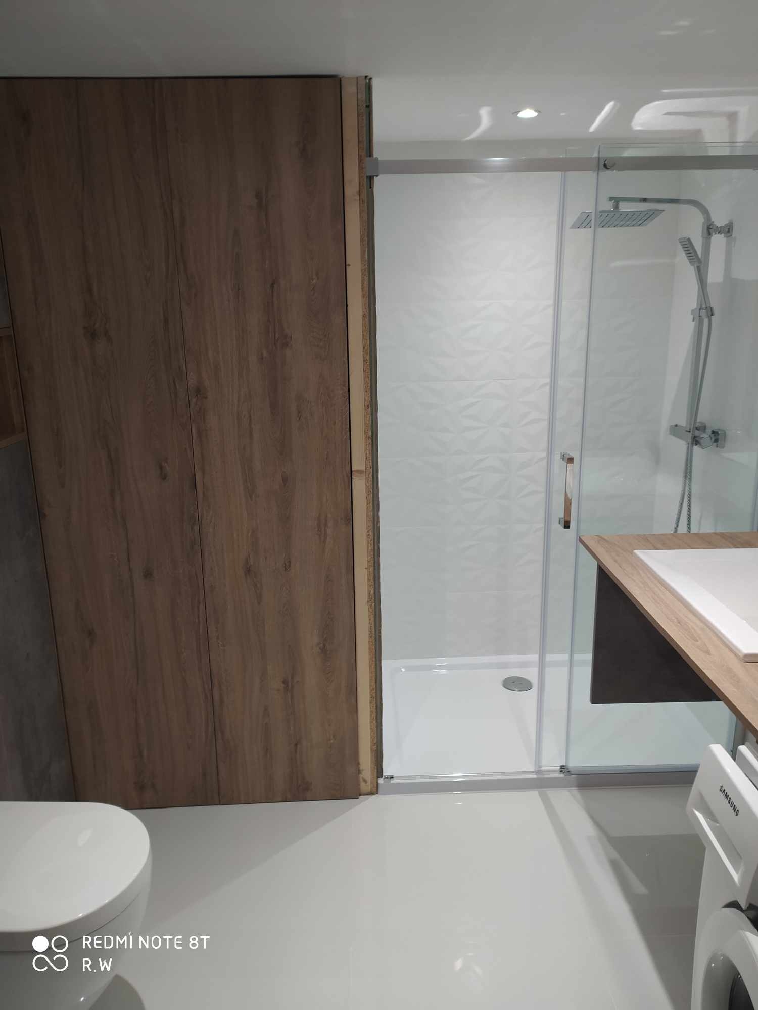 Wykończona łazienka z kabiną prysznicową, umywalką z drewnianym blatem, zabudową w kolorze drewna i podwieszaną toaletą, widoczna pralka.